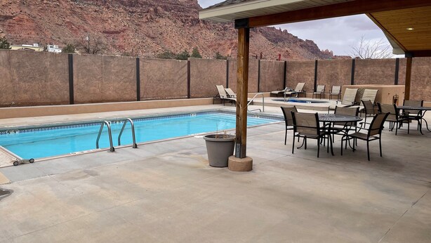 Área de piscina com espreguiçadeiras e mesas, um alpendre coberto e vista para formações rochosas avermelhadas.