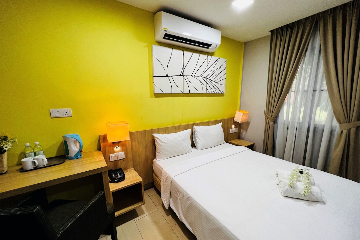 A hotel room with a bed, a desk with a lamp, an air conditioner, and a wall-mounted artwork.