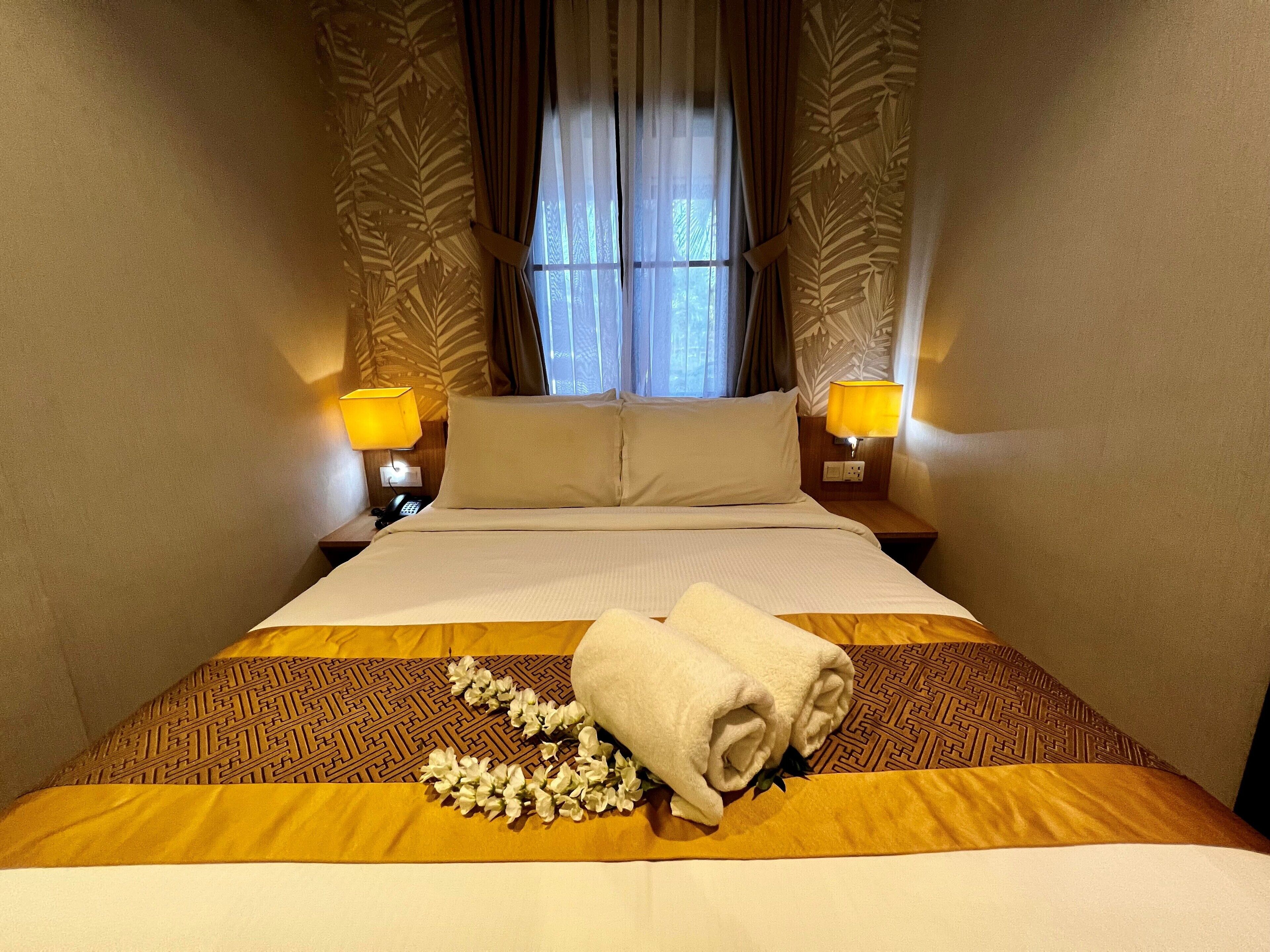 A hotel room with a large bed, two bedside lamps, a window with curtains, and a floral arrangement on the bed.