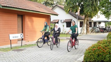 Un camino pavimentado con tres ciclistas, un cartel que dice "SURAU" y un edificio con techo de tejas.