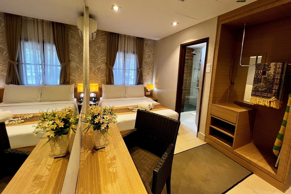 Ein Hotelzimmer mit einem großen Bett, einem Holzespeisetisch mit Blumen, einem Stuhl, einem kleinen Tisch mit Lampe und einer Duschkabine.