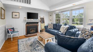 A spacious living room with a fireplace, a sofa set, a television, and a large window offering a view of the outdoors. - Turtle Cove Cottage: Steps to Long Pond Beach (South Yarmouth)