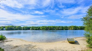 A lakeside view with a boat on the sand, calm water, and a forested shoreline. - Turtle Cove Cottage: Steps to Long Pond Beach (South Yarmouth)