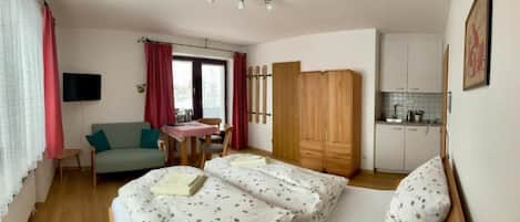Una habitación con cama, sofá, mesa, televisor y zona de cocina.