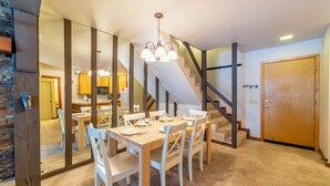 Ein Essbereich mit einem Holztisch, gedeckt für vier Personen, weißen Stühlen und einem Kronleuchter. Im Hintergrund ist eine Treppe mit Holzwandung zu sehen.