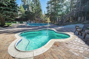 A swimming pool with a unique shape surrounded by a brick patio, lounge chairs, and a wooden deck.