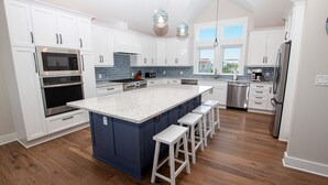 A modern kitchen with white cabinets, a large island with bar stools, stainless steel appliances, and a view of the outside through large windows. - Oceanside, Private Pool, ELEV, Hot Tub Rec Room, yard, SUN-SUN, WW865-This One's for You! (Corolla)