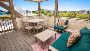 A covered outdoor deck with a wooden table and chairs, a sofa with a patterned cushion, and a view of houses and greenery. - Oceanside, Private Pool, ELEV, Hot Tub Rec Room, yard, SUN-SUN, WW865-This One's for You! (Corolla)