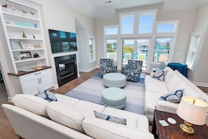 A living room with a fireplace, white sofas, ottomans, and a TV. - Oceanside, Private Pool, ELEV, Hot Tub Rec Room, yard, SUN-SUN, WW865-This One's for You! (Corolla)