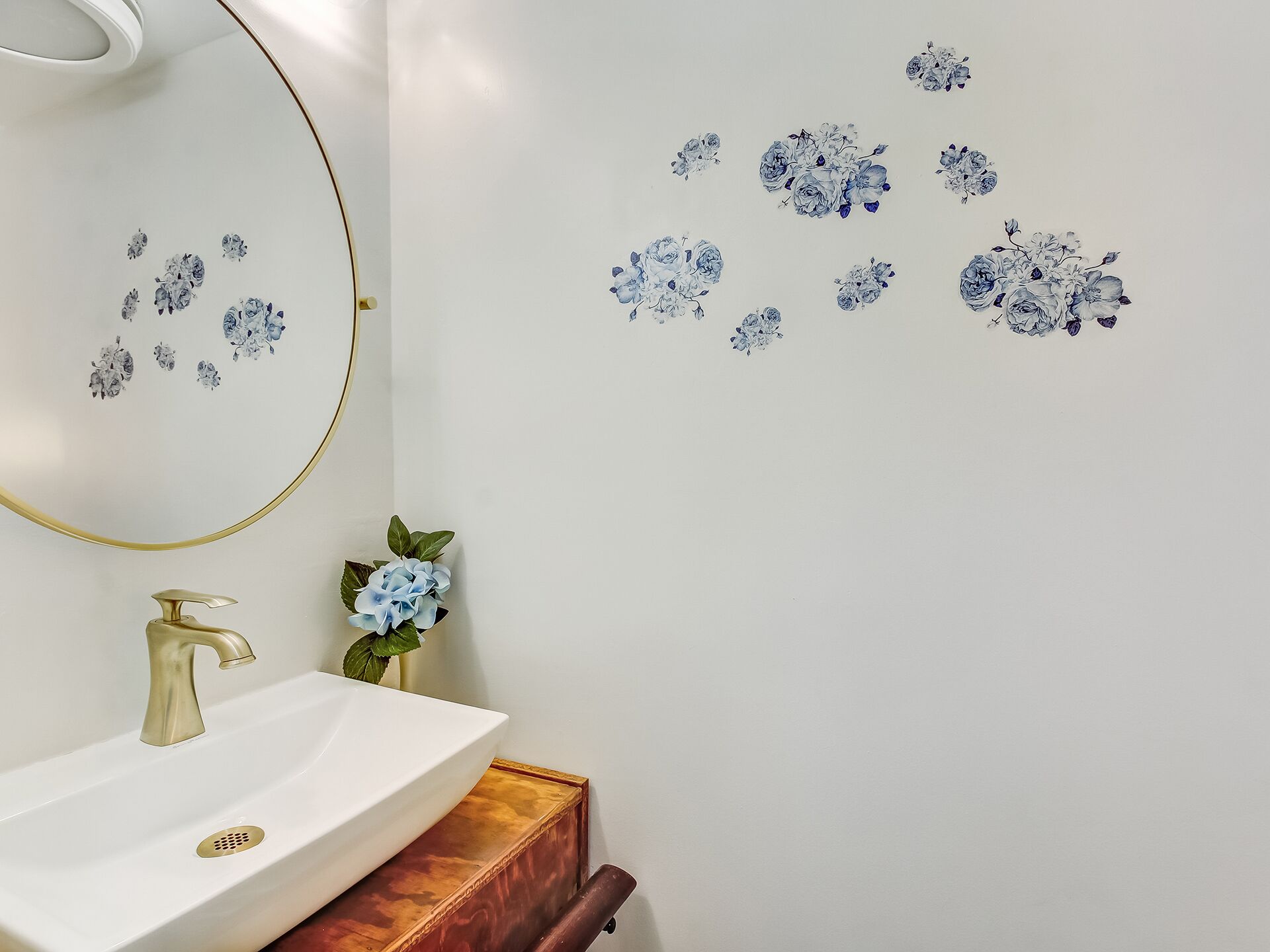 A bathroom with a white sink, gold faucet, and a mirror with a gold frame. There are blue floral decals on the wall and a small potted plant on the counter.