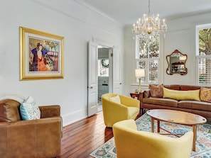 A living room with a brown leather sofa, yellow armchairs, a round wooden coffee table, and a chandelier. - Heated Pool Access, Exquisite Historic Home (Savannah)