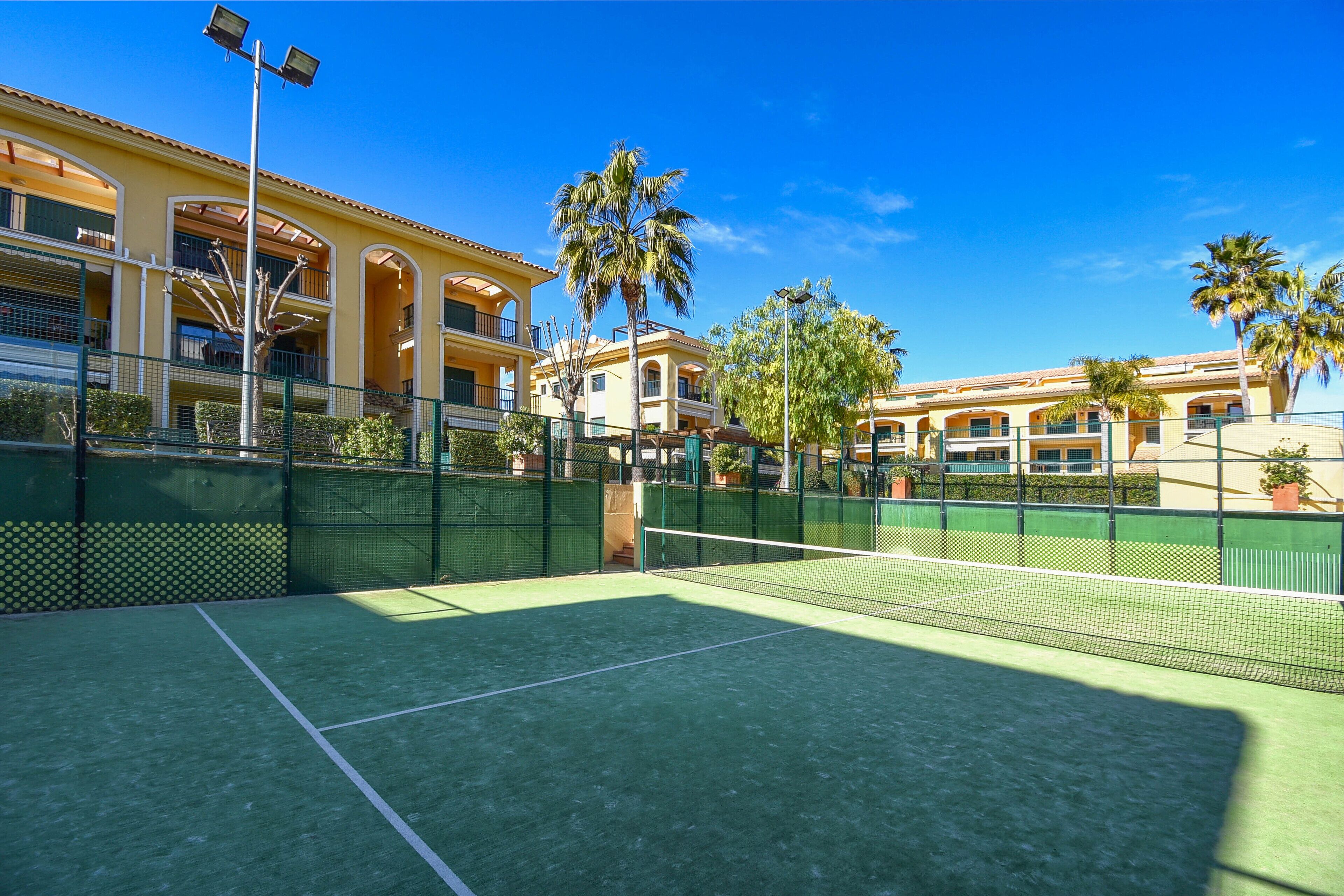 Un court de tennis avec un filet, entouré d’une clôture verte, et situé à côté d’un bâtiment doté de balcons et de palmiers.