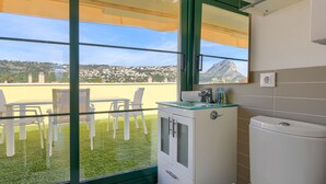 A bathroom with a toilet, a sink, and a mirror. There is a window with a view of a green lawn and buildings. - Sorolla Sol - Javea, Costa Blanca (Javea)
