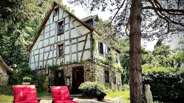 Una casa tradicional de entramado de madera con base de piedra, una puerta roja y un pequeño jardín con dos sillas rojas.