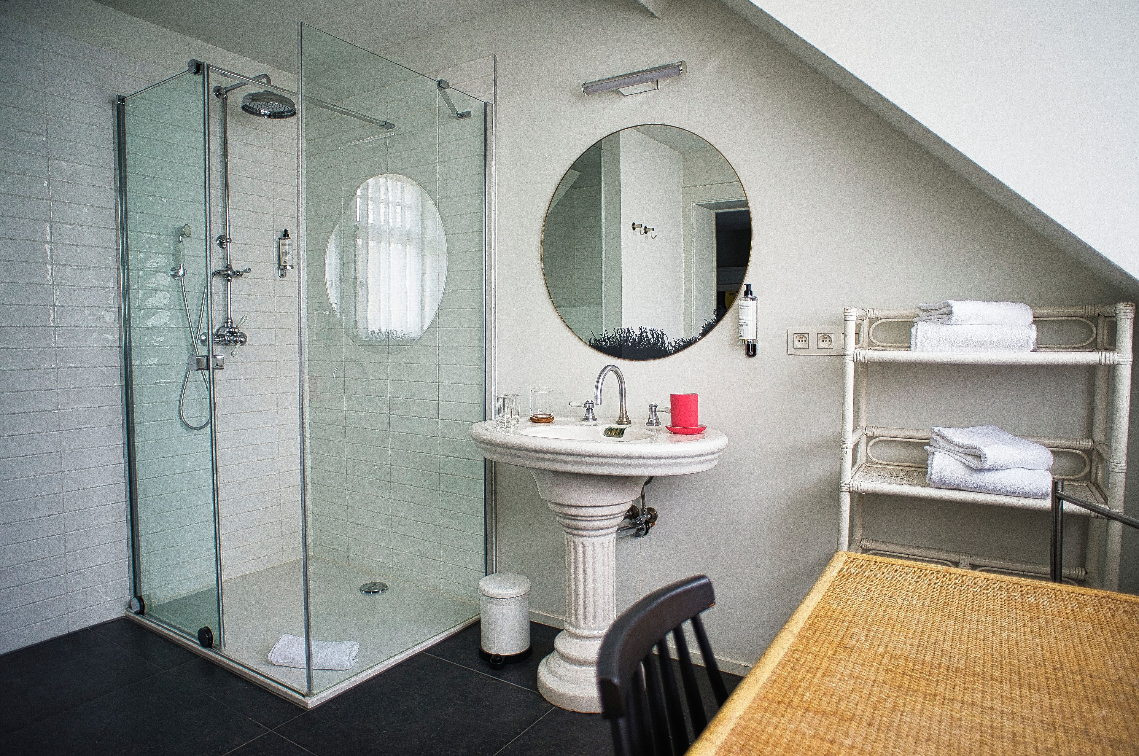 Un baño con ducha de mampara de vidrio, lavamanos pedestal, espejo redondo y una pequeña mesa con una silla.