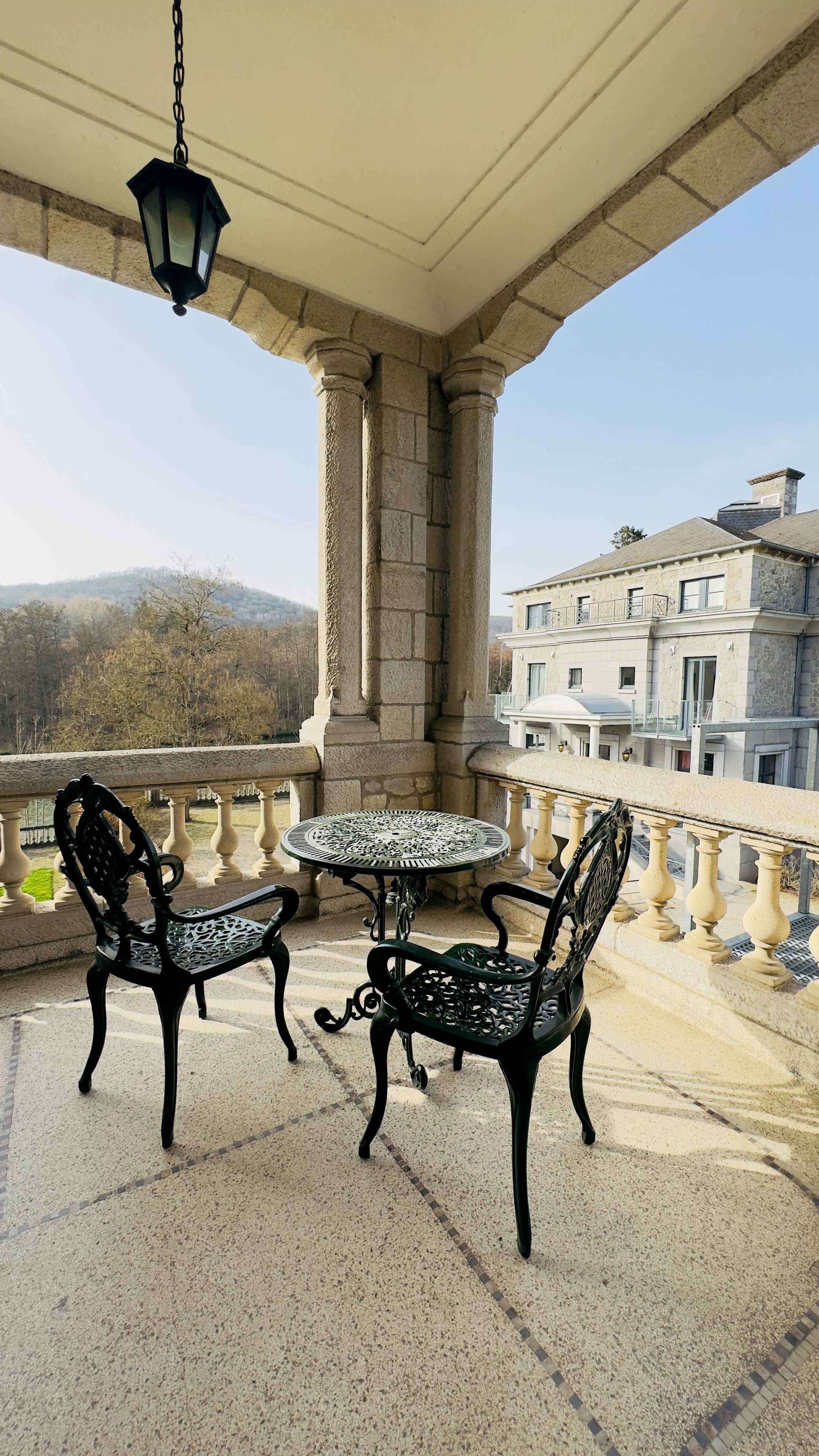A balcony with two chairs and a table, stone columns, and a view of buildings and trees.
