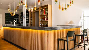 A modern bar area with a wooden bar counter, hanging pendant lights, and a seating arrangement with black chairs.