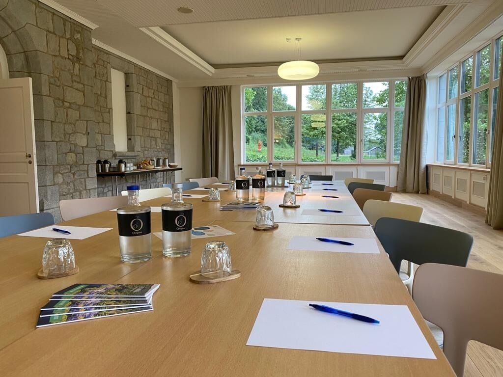 Une salle de conférence avec une longue table, des chaises et un mur en pierre. On peut y voir des bouteilles en verre, des papiers et des stylos sur la table.