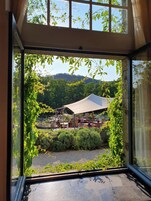 Vue depuis une pièce dotée de grandes fenêtres, donnant sur un jardin avec un espace salon abrité et un paysage montagneux en arrière-plan.