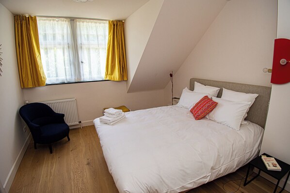 A bedroom with a large bed, a chair, a radiator, and a window with curtains. - Les Sorbiers (Hastiere)