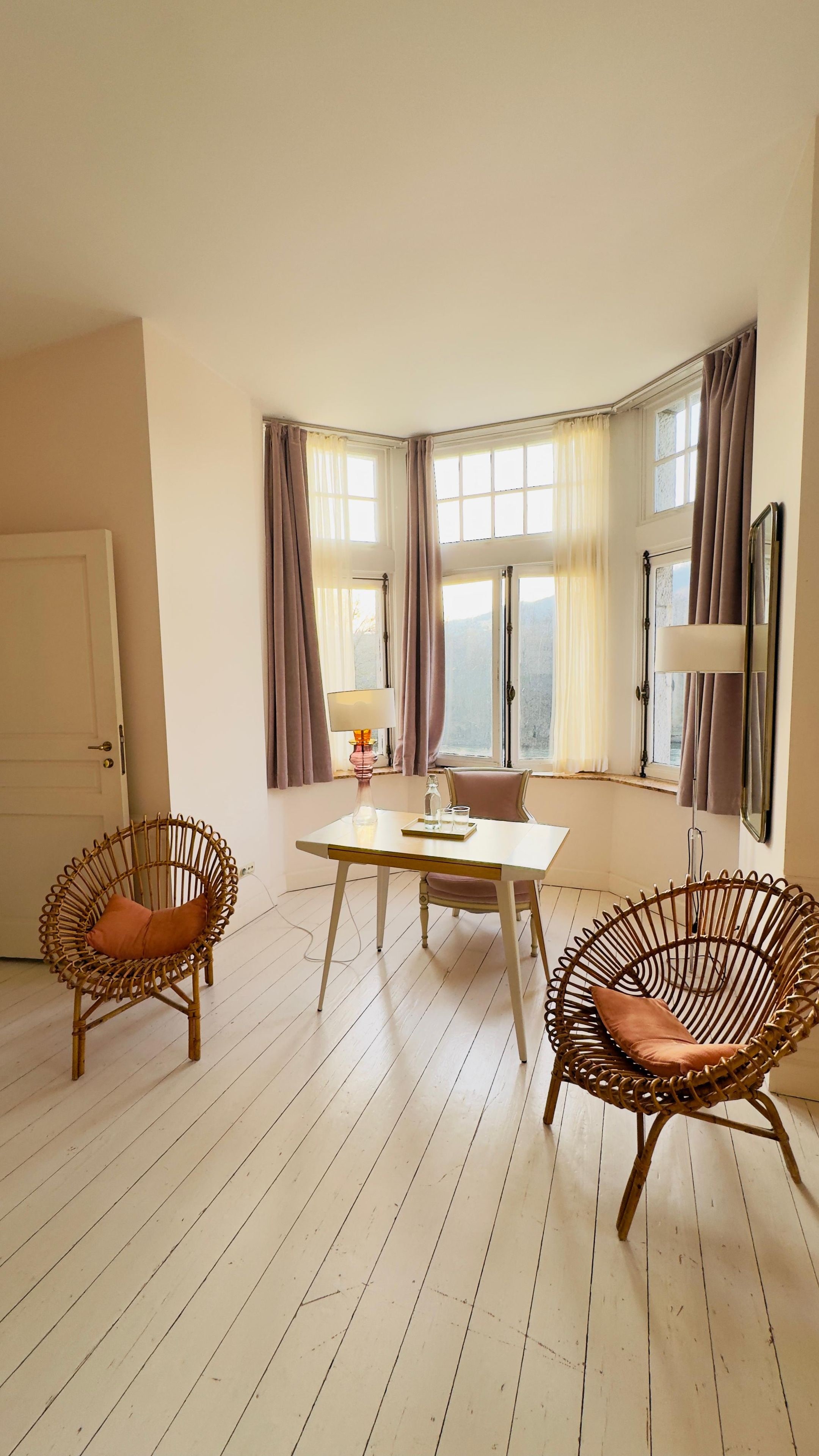 A room with a dining table, two wicker chairs, a lamp, and a window with curtains.