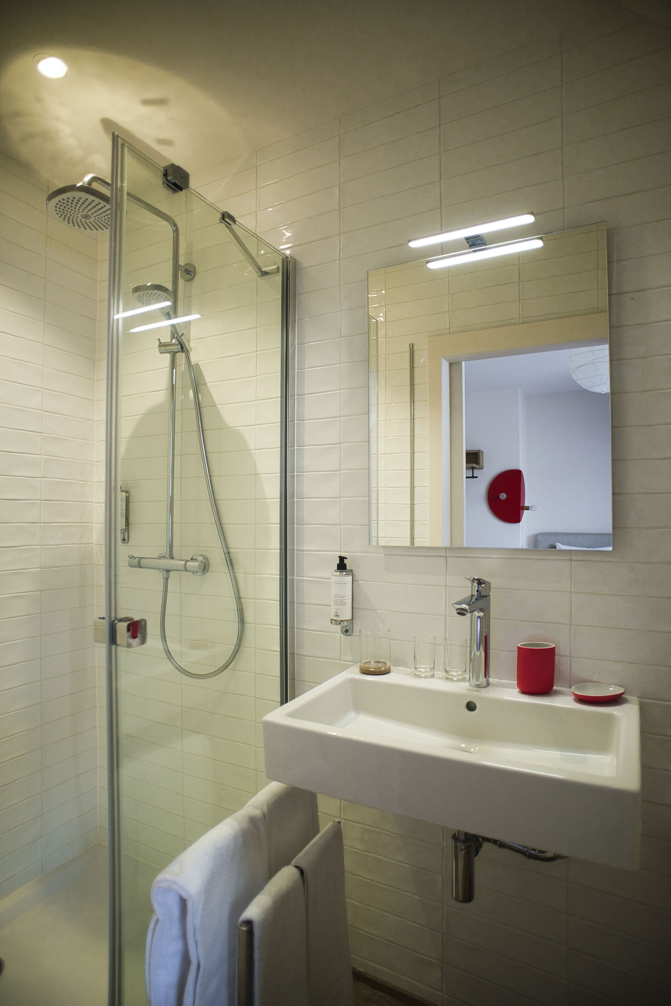 Un baño moderno con ducha de vidrio, un lavamanos blanco y un espejo con un detalle rojo.