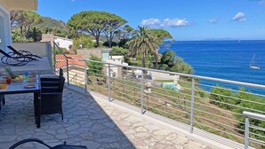 A terrace with a table set for dining, overlooking a coastal area with a clear view of the sea and boats.