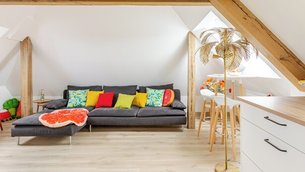 A modern living room with a gray sectional sofa, colorful pillows, a wooden ceiling, and a white dining area with chairs. - Maison 1934 bnb (Colmar)