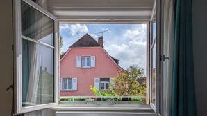 A window with a view of a pink house with white shutters and a chimney. - Maison 1934 bnb (Colmar)