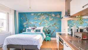 A modern bedroom with a large bed, a blue wall mural, a kitchen area with a toaster, and a window with curtains. - Maison 1934 bnb (Colmar)