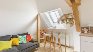 A modern loft apartment with a sloped ceiling, a dining area with bar stools, a gray sofa with colorful cushions, and a wooden-framed window. - Maison 1934 bnb (Colmar)