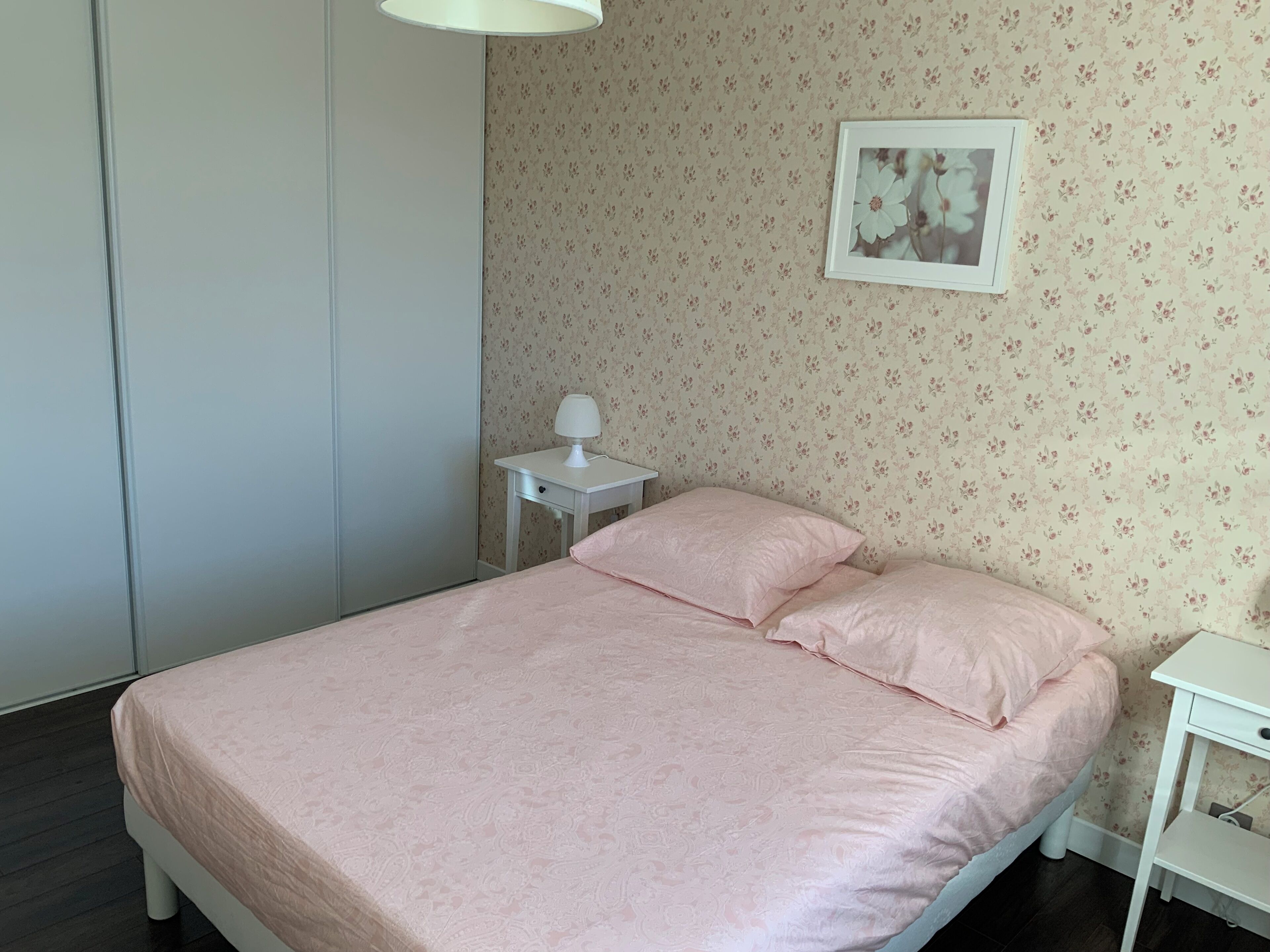 Ein ordentlich gemachtes Bett mit rosa Bettdecke und Kissen, ein kleiner Nachttisch mit Lampe sowie eine Wand mit floralem Wandpapier.