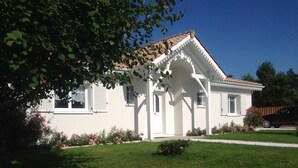 Une maison blanche avec un toit en tuiles, un porche à l’avant et un petit jardin orné de fleurs roses.