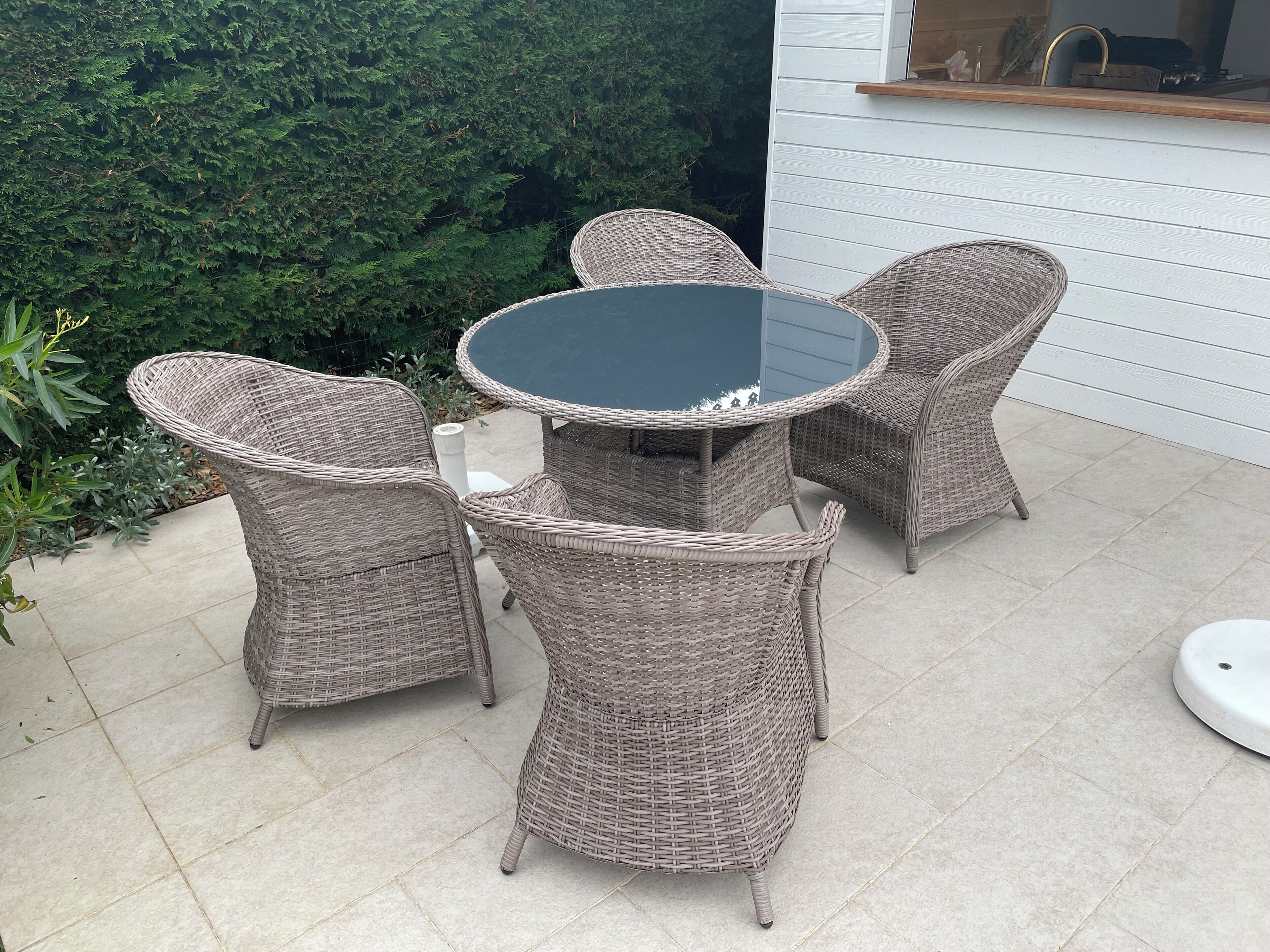 A wicker outdoor dining set with a glass table and four chairs, positioned on a tiled patio with a white wall and greenery in the background.