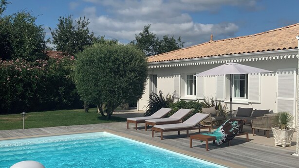 Une piscine dotée de chaises longues et de bouées gonflables, entourée d’un jardin aménagé et d’un bâtiment dont le toit est recouvert de tuiles.