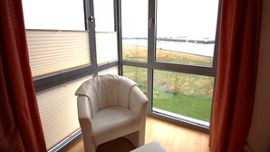 Ein Zimmer mit großem Fenster, einem weißen Stuhl und Blick auf eine Wiese sowie eine Brücke.