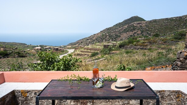 Ein Tisch mit einer Flasche, Gläsern und einem Hut auf einer Terrasse mit Blick auf eine malerische Landschaft.