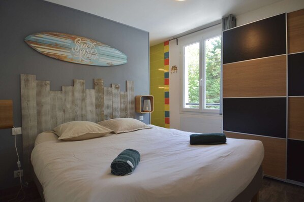 Une chambre avec un lit, une planche de surf accrochée au mur, un mur aux couleurs vives et une fenêtre donnant sur un paysage verdoyant.