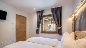 Une chambre avec une porte en bois, une fenêtre avec des rideaux, un lit avec une literie blanche et un téléviseur fixé au mur.