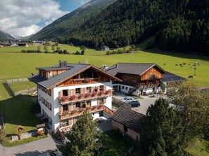 Ein mehrgeschossiges Gebäude mit Balkonen, einem Parkplatz und Blick auf ein grünes Feld und bewaldete Berge.