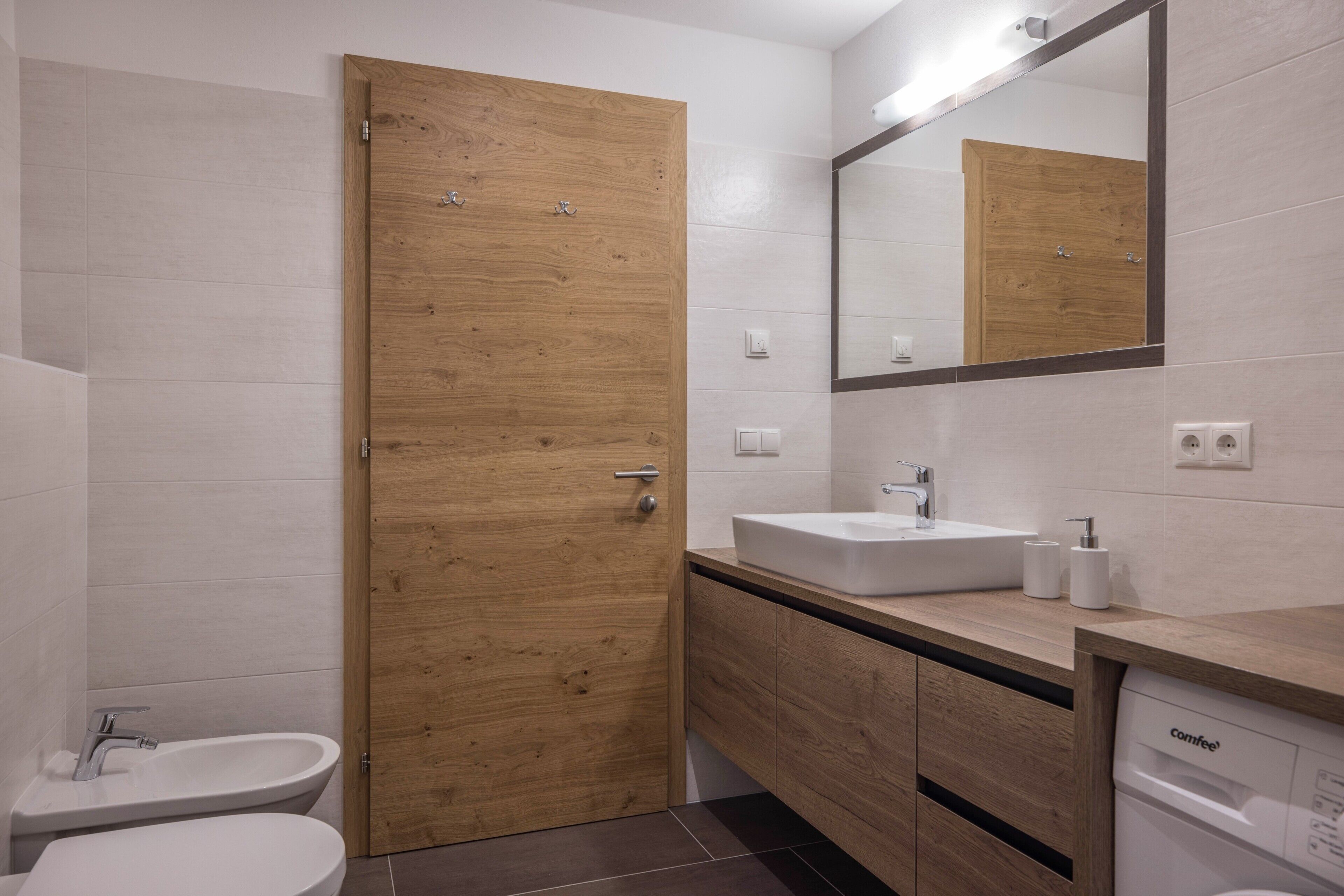 Une salle de bain moderne avec une porte en bois, un lavabo blanc, des toilettes et un lave-vaisselle encastré.
