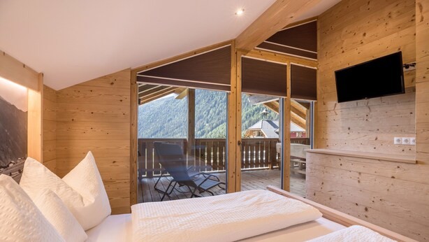 Interior de madera con una cama, un televisor montado en la pared y un balcón con vistas a las montañas.