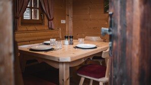 A wooden cabin interior with a dining table set for two, a window with curtains, and a door. - Cottage 'Lechnerhütte Fane Alm' with Mountain View, Wi-Fi & Terrace (Vals)