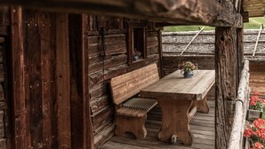 A rustic wooden cabin with a wooden bench and table, a small window with a flower vase, and a wooden deck with a railing. - Cottage 'Lechnerhütte Fane Alm' with Mountain View, Wi-Fi & Terrace (Vals)