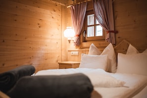 Una habitación de cabaña de madera con una cama, almohadas, una mesita de noche, una lámpara y cortinas a cuadros.