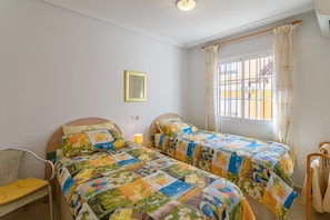 A bedroom with two beds, a chair, a bedside table, a window with curtains, and a wall-mounted light fixture.