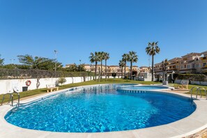 A clear blue swimming pool with a curved edge, surrounded by palm trees and a building with a tiled roof in the background. - Holiday Home 'Casa Susan' with Pool, Wi-Fi & Garden (Torrevieja)