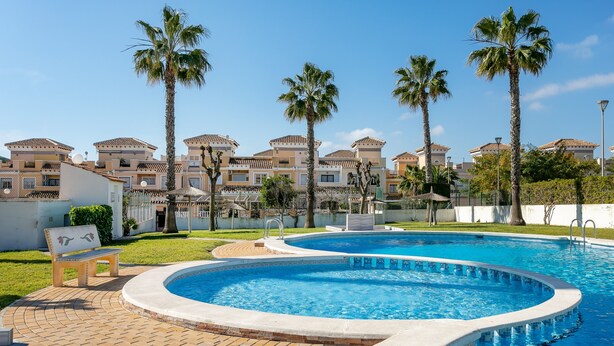 Une piscine avec une terrasse carrelée, des palmiers et, au fond, un bâtiment dont le toit est également recouvert de tuiles.