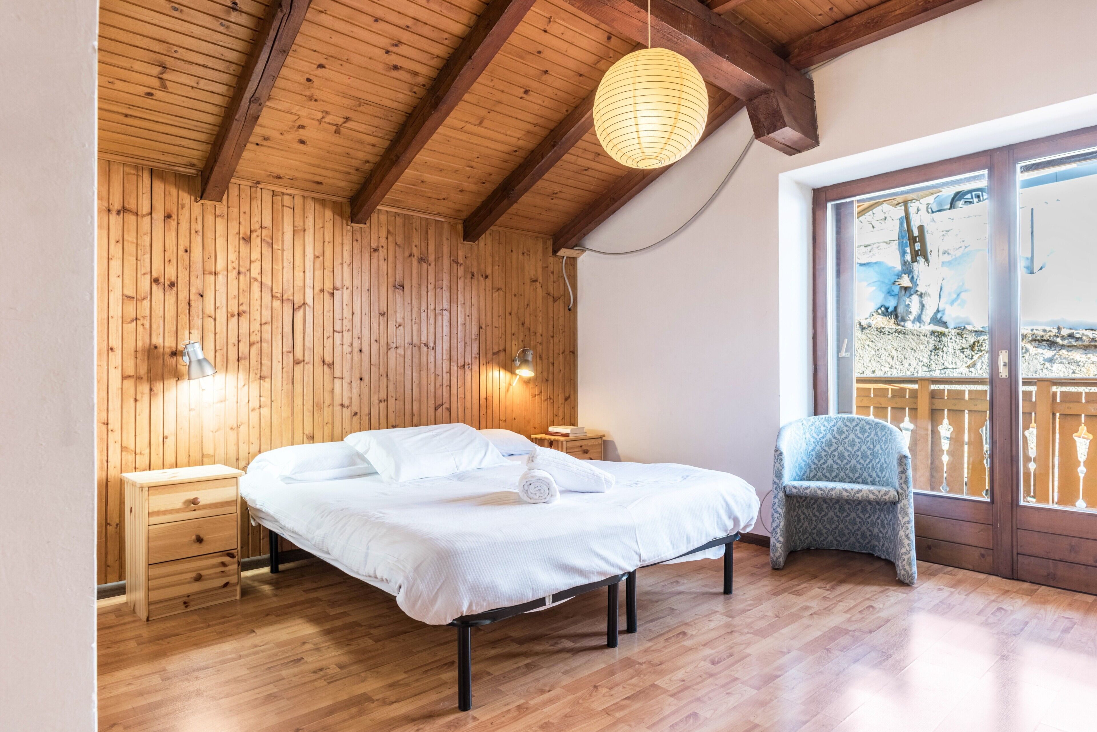Habitación con paneles de madera, una cama, mesitas de noche, una silla y vistas a través de un ventanal.