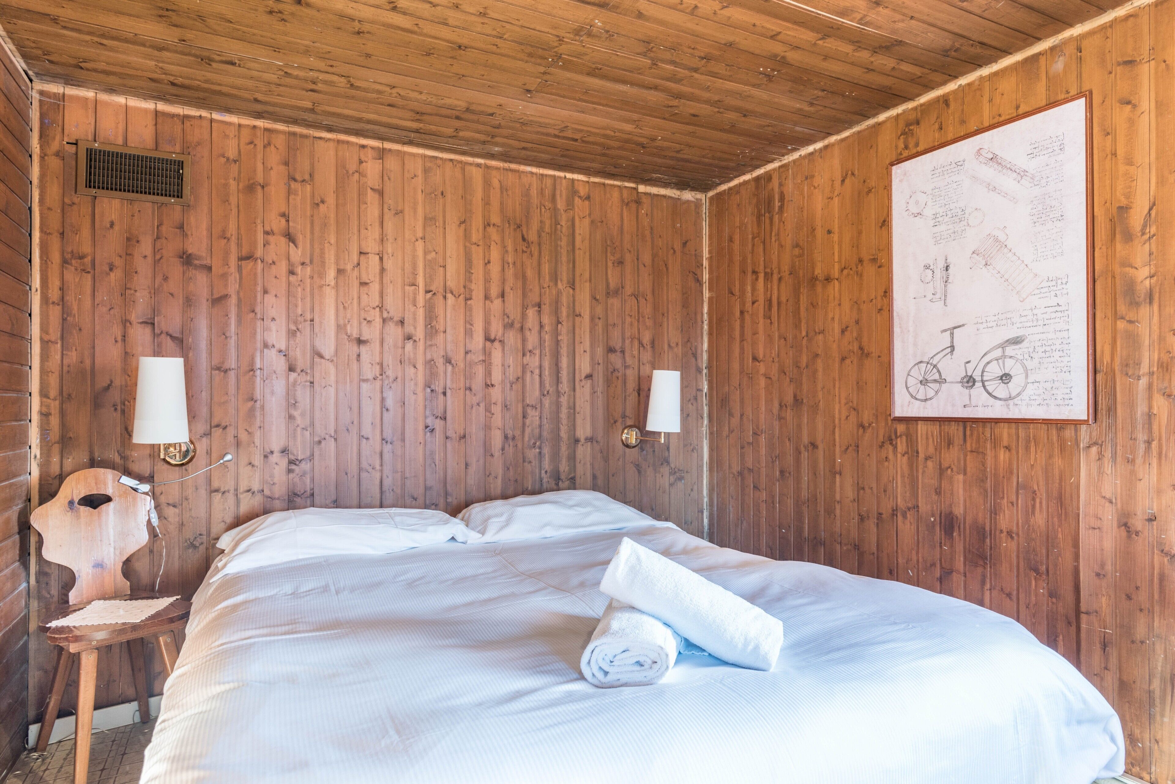 Una stanza con rivestimento in legno, un letto, due lampade a parete, una sedia e un diagramma incorniciato appeso al muro.